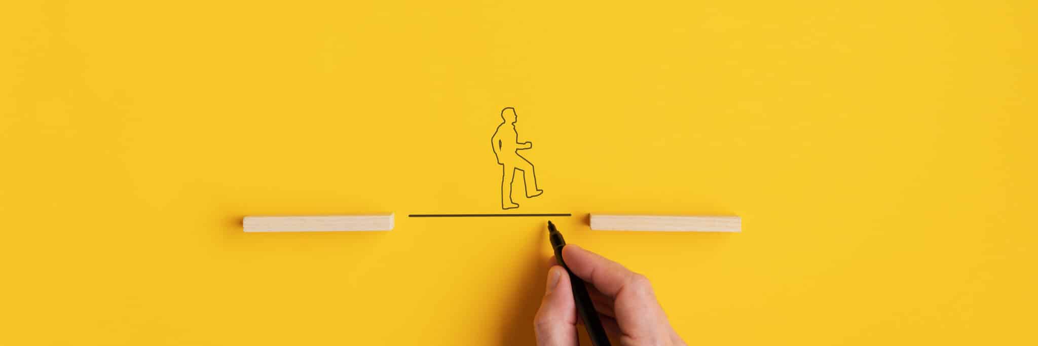 Wide view image of male hand drawing a line between two wooden pegs for a silhouette of a man to walk across. Over yellow background with copy space.