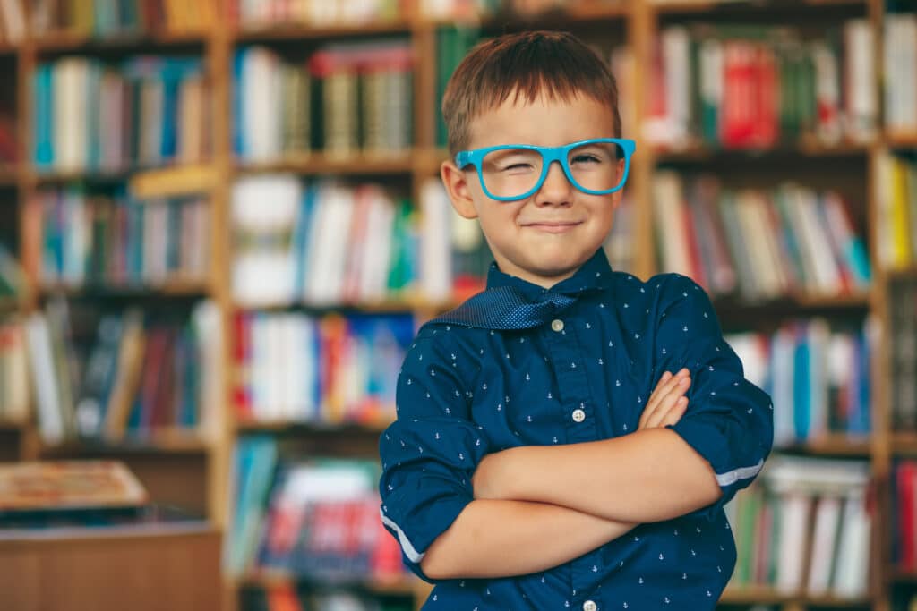 Smiling boy winking with blue glasses frame and blue tie on the shoulder on a book background. Pupil holds cross hands against multi colored bookshelf in library. Education, Knowledge, School.