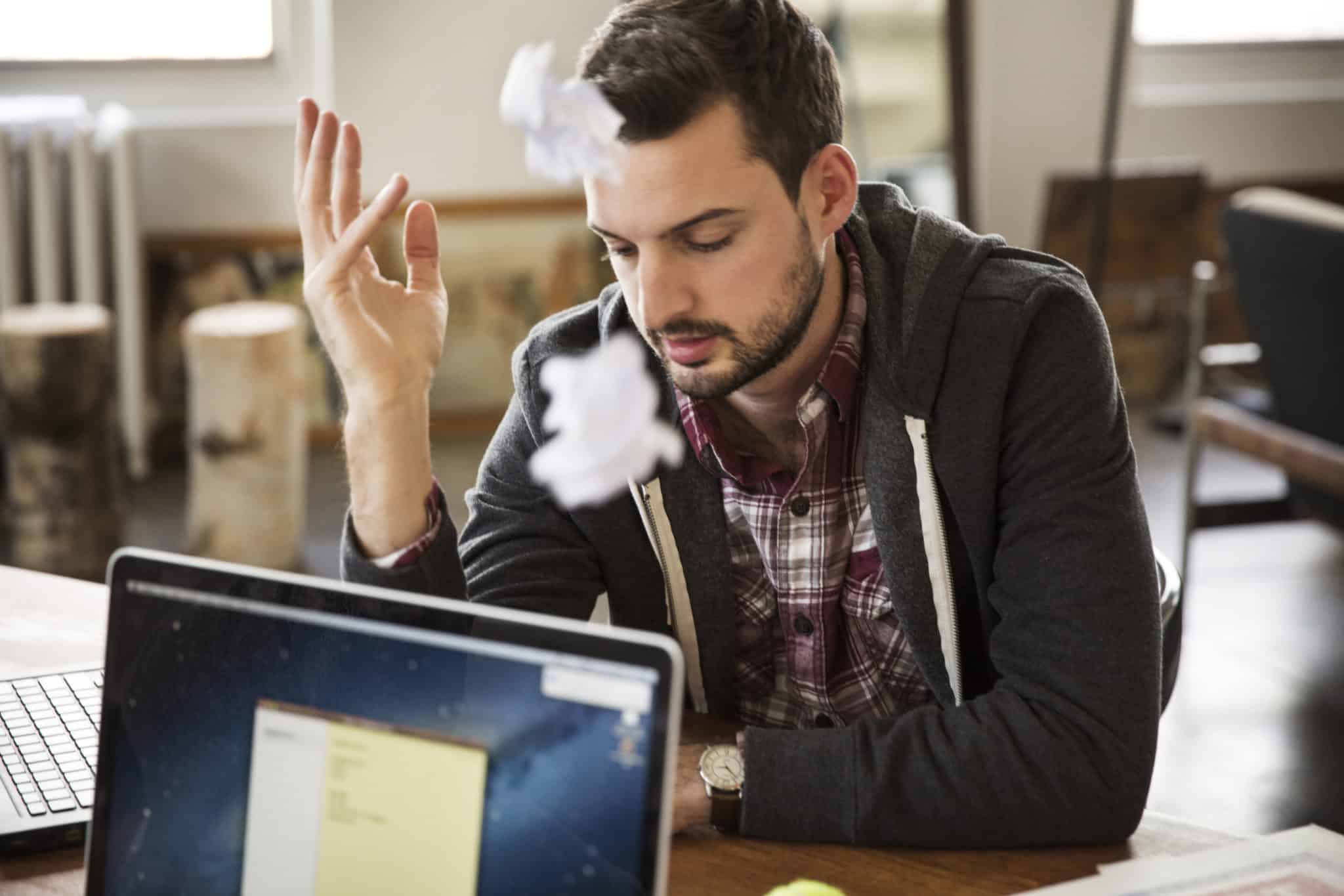 Frustrated Worker Sitting By Laptop On Table With Crumbled Paper In Mid-air At Office