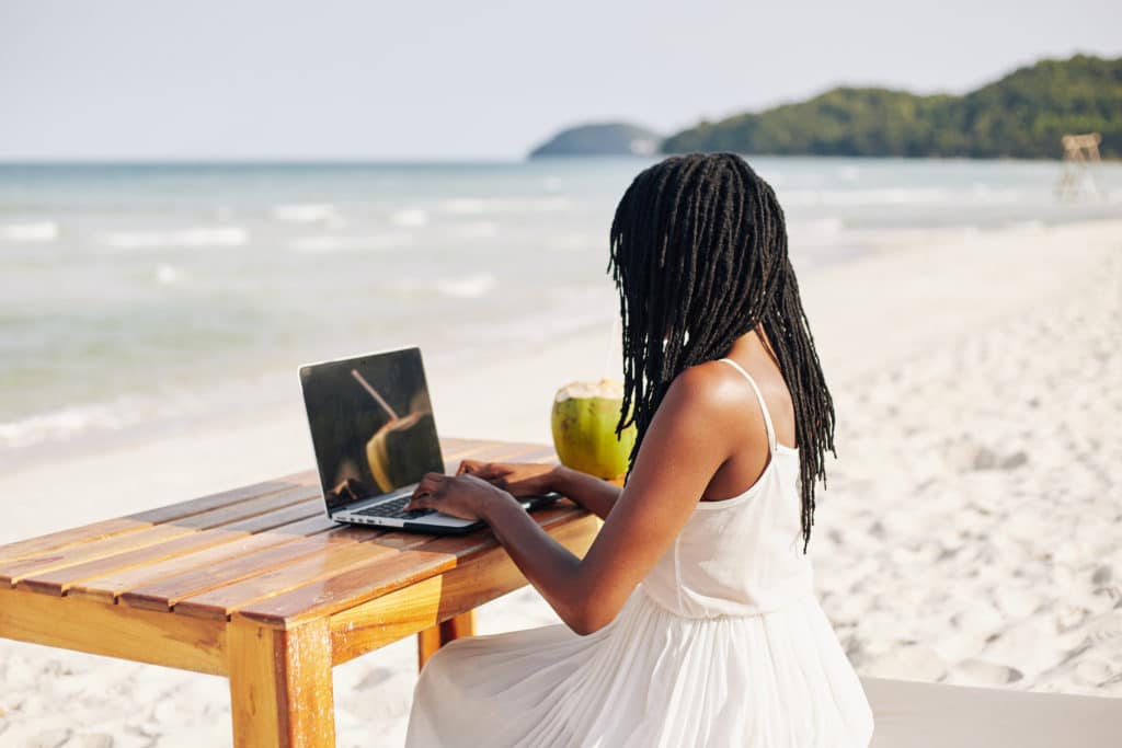 Young Black woman with dreadlocks sitting at wooden table on beach and working on laptop