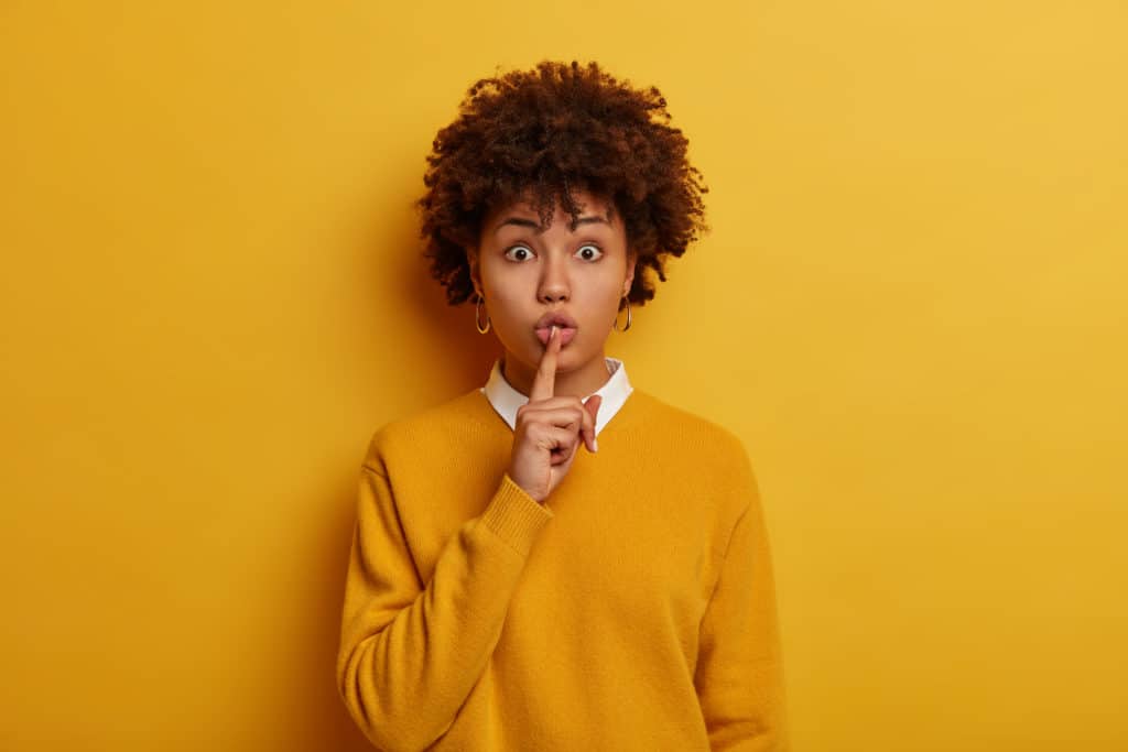 Shush, be quiet. Mysterious wondered Afro American woman makes hush sign, asks be silent, tells very secret information or story to interlocutor, wears bright yellow sweater, spreads rumors.