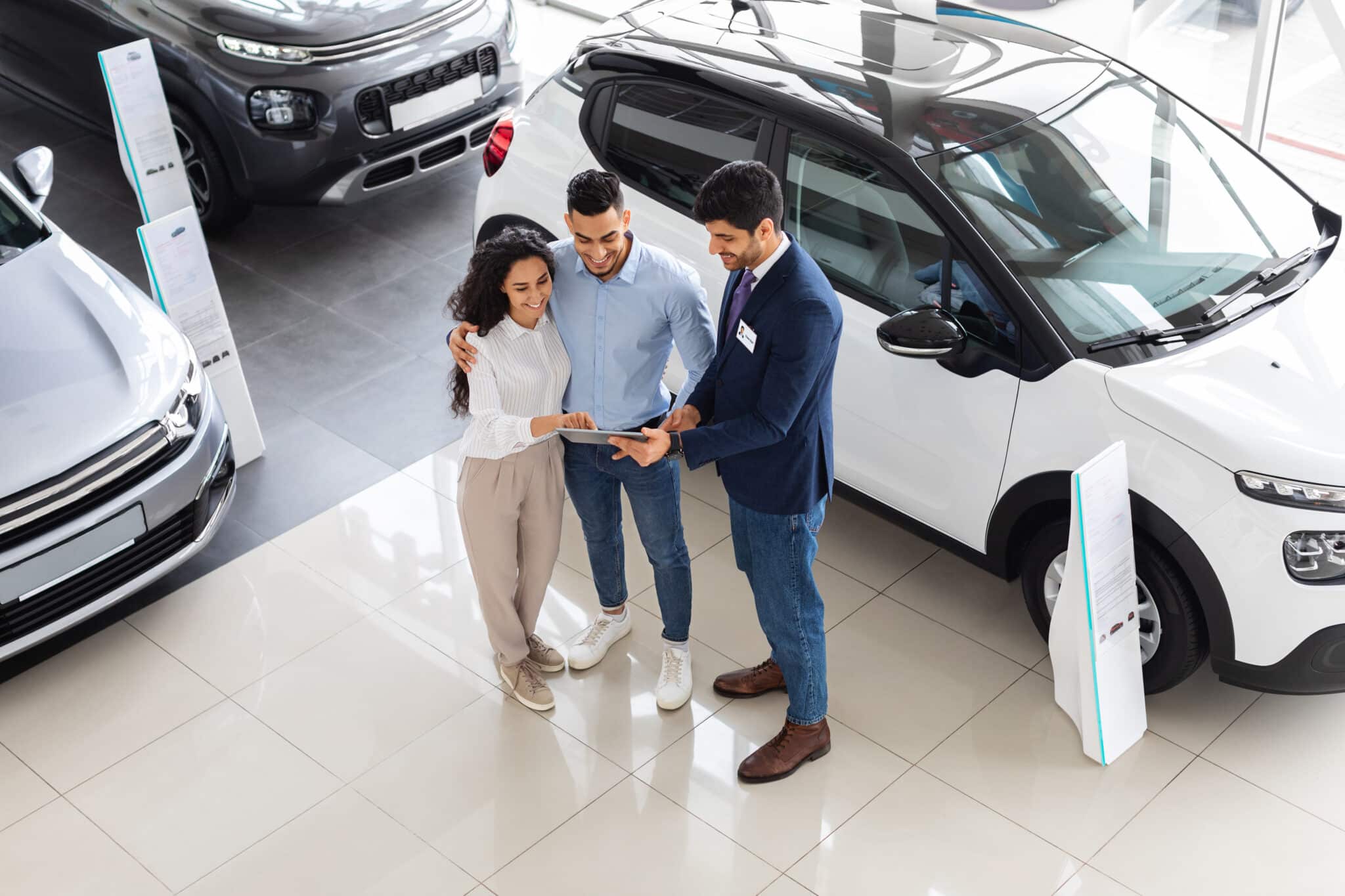 Top view of cheerful middle-eastern young couple having conversation with good-looking sales assistant at auto showroom, checking papers or car specification, full length photo
