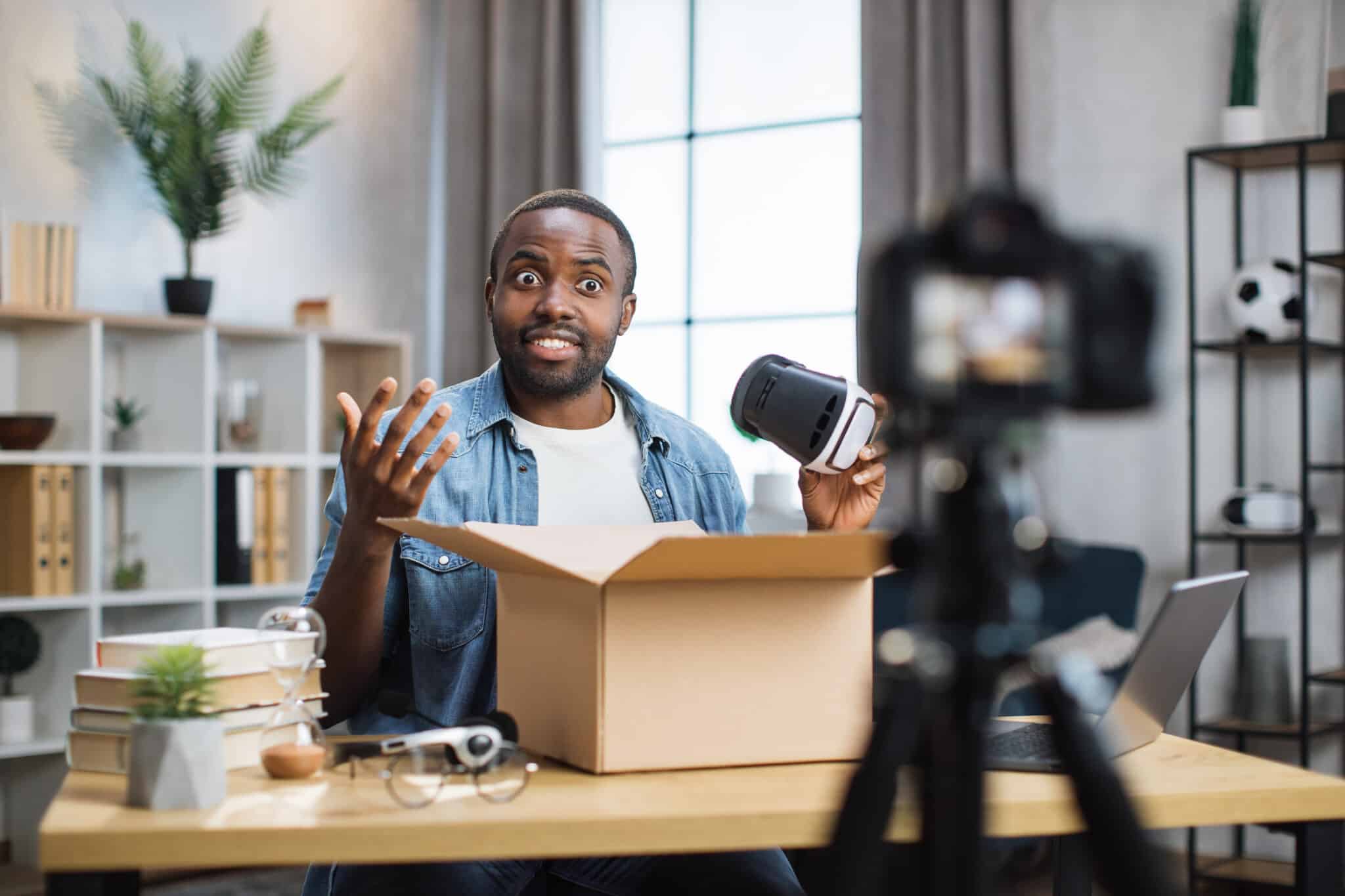 African american man in casual wear opening parcel box and recording video at home. Male influencer sitting at table with VR glasses in hands and talking on camera.